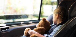 Ein Mädchen sitzt in einem Kindersitz im Auto und schläft. Sie hält einen Teddybären dabei.