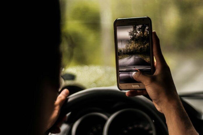 Eine Person fährt auf einer Landstraße im Auto. Sie hält ein Smartphone in der rechten Hand, der Daumen am Auslöser der Kamera. Die linke Hand ruht am Steuer.